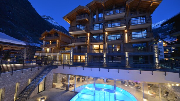 A multi-story hotel with balconies, a swimming pool, and a view of snow-capped mountains. - Alpenhotel Fleurs de Zermatt (Zermatt)