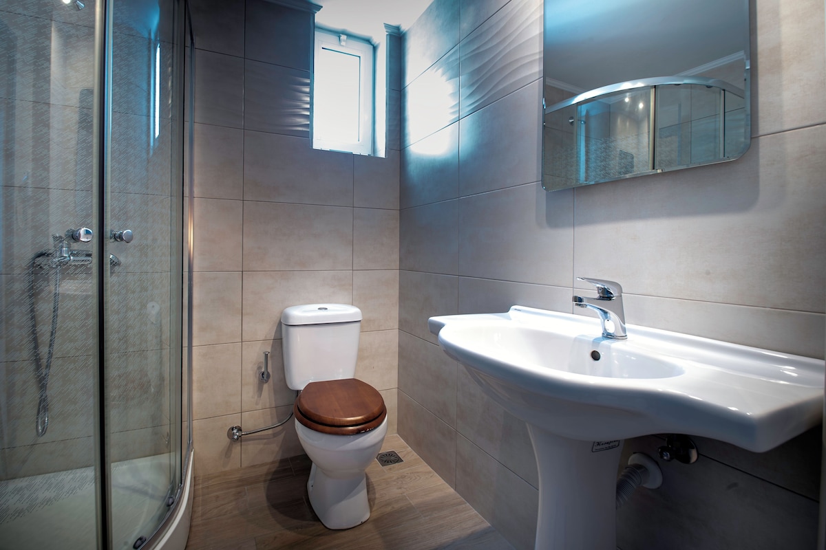 A modern bathroom with a glass-enclosed shower, a white toilet with a wooden seat, and a white sink with a mirror above it.