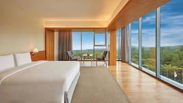 A spacious hotel room with a large bed, a small table with chairs, and a view of the outdoors through large windows.