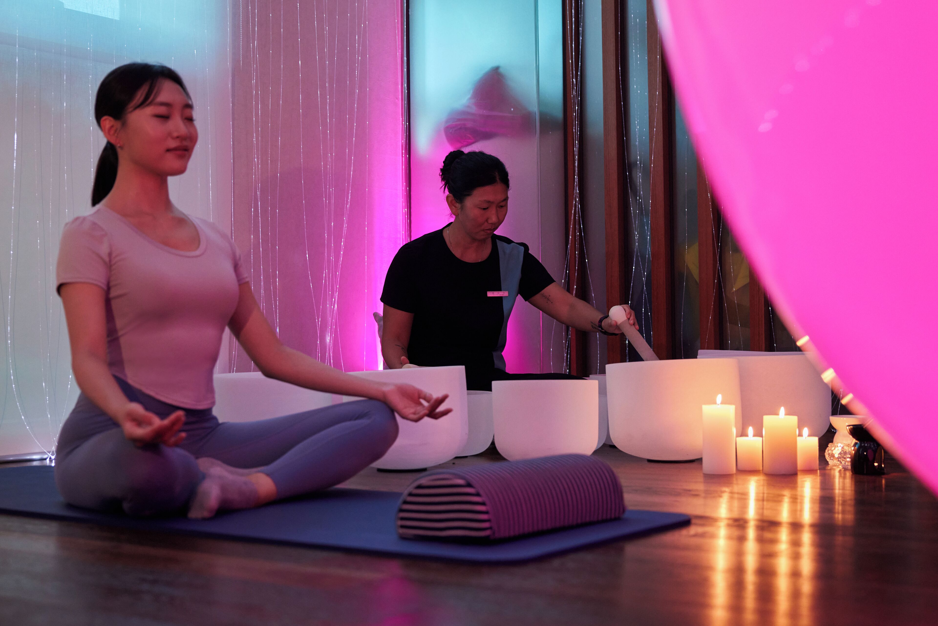 Dos personas meditando sobre esterillas de yoga en una habitación con velas y un globo rosa grande.