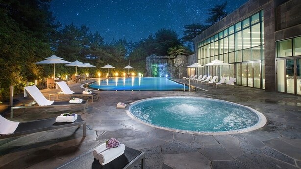 Un área de piscina nocturna con sillones de descanso y sombrillas, un jacuzzi y un edificio con amplias ventanas.