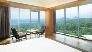Amplia habitación con grandes ventanales que ofrecen vistas a un paisaje boscoso y montañas. Se puede observar una cama, un sillón y una pequeña mesa.
