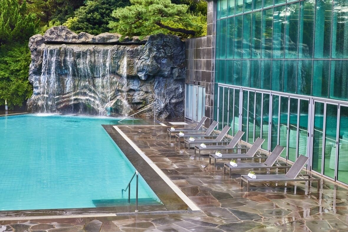 En pool med ett vattenfall, solstolar och en byggnad med glasfasad som vetter mot poolen.