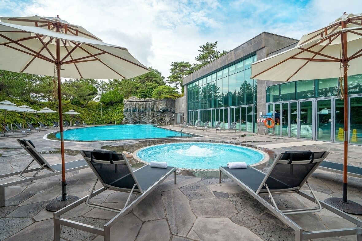 A swimming pool with lounge chairs and umbrellas, surrounded by a stone patio and a modern building with large windows.