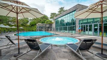 Ein Pool mit Liegestühlen und Sonnenschirmen, umgeben von einer Steinterrasse und einem modernen Gebäude mit großen Fenstern.