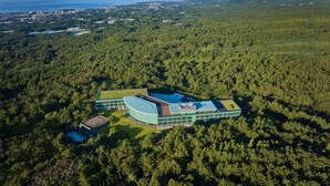 Vista aérea de un edificio moderno con techo curvo, rodeado de un bosque frondoso, con vistas al mar y colinas distantes.