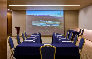 Una sala de conferencias con una pantalla grande mostrando una imagen de hotel, varias mesas con sillas y un atril.