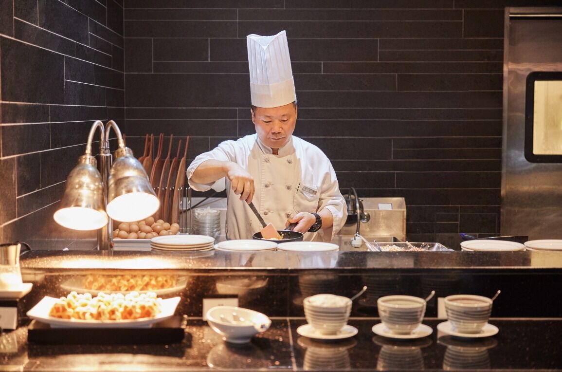 A chef preparing food in a kitchen with a black tiled backsplash and stainless steel appliances.