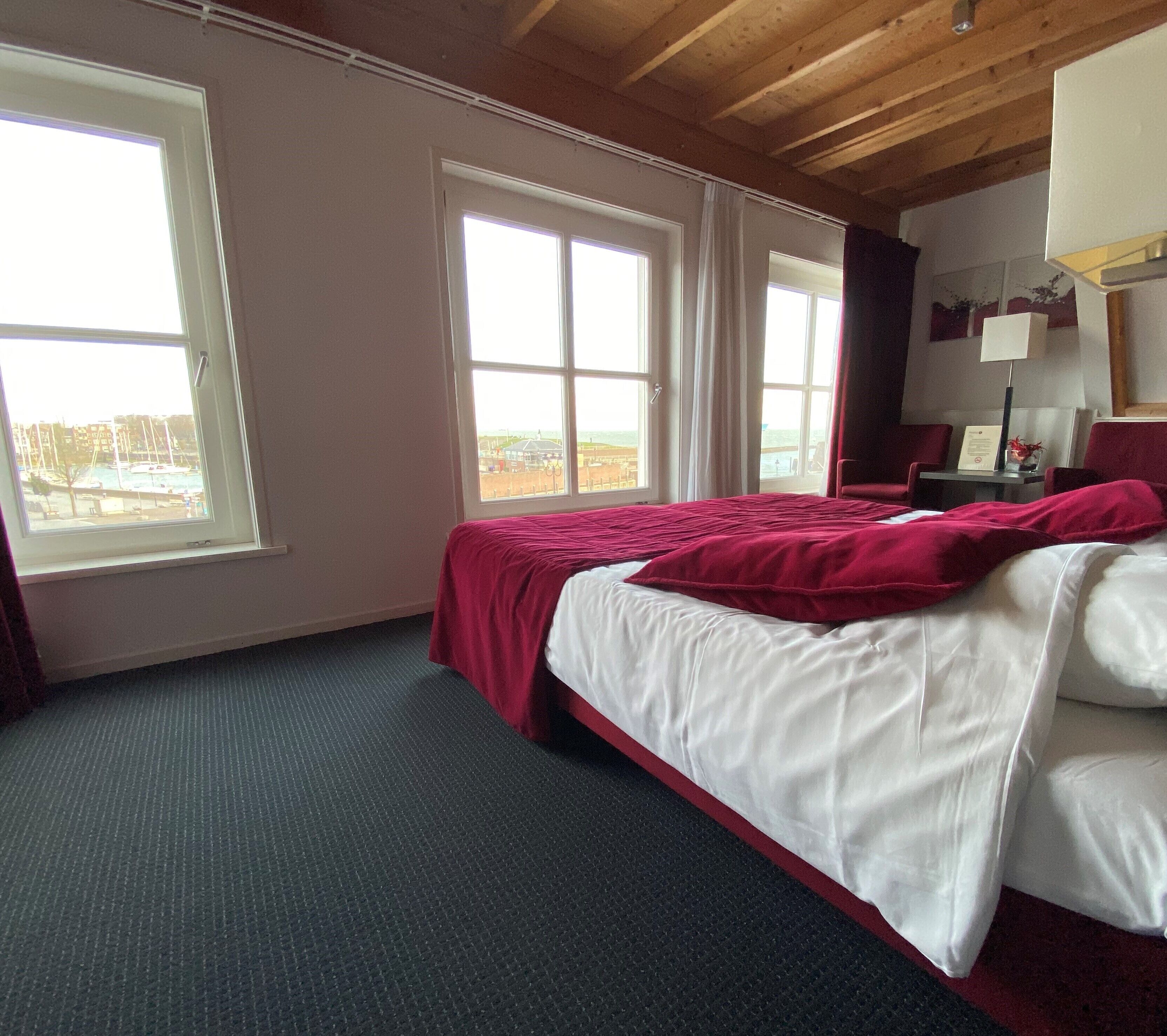 Ein Hotelzimmer mit einem großen Bett, zwei Fenstern, einem Schreibtisch mit Lampe und Meerblick.