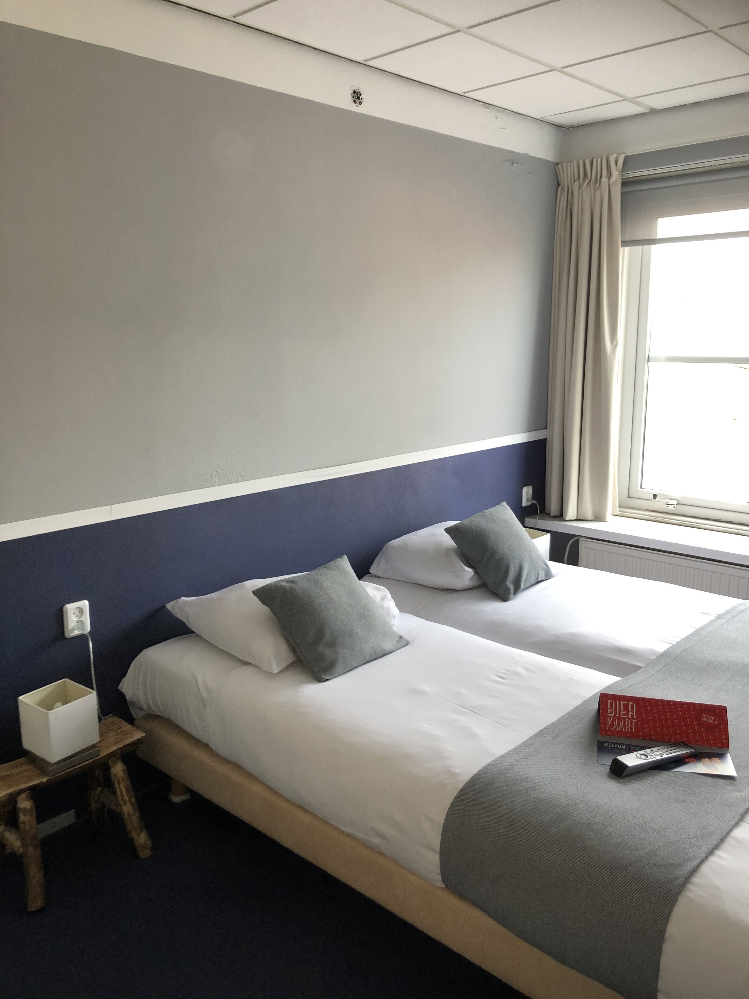 A hotel room with two beds, a bedside table, a lamp, a window with curtains, and a book on the bed.