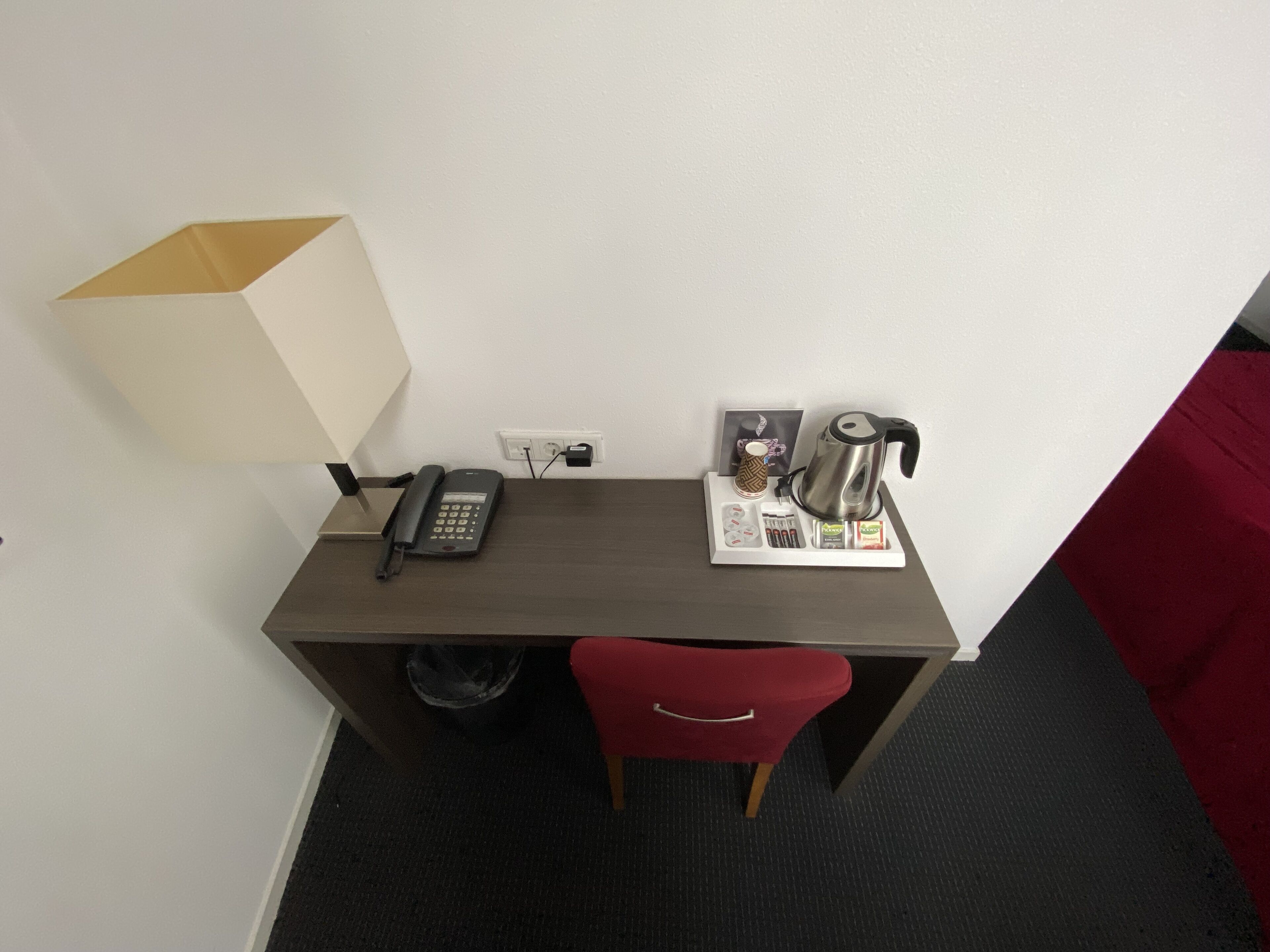A hotel room with a desk, a telephone, a kettle, and a small table with a cup and a book.