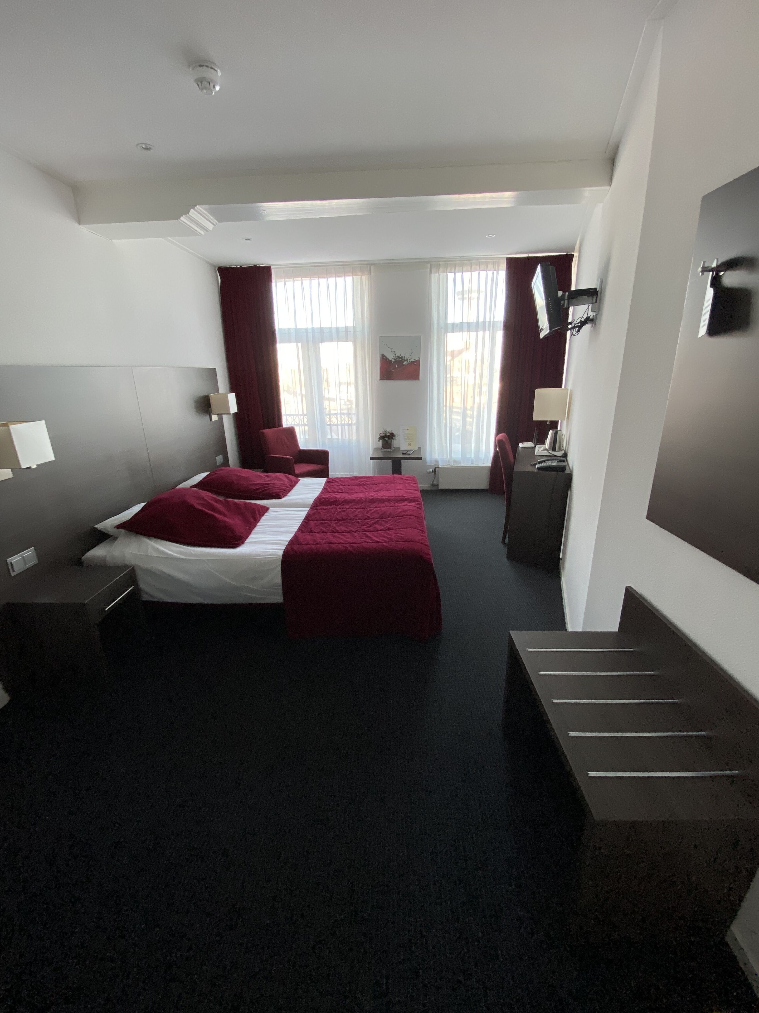 Una habitación de hotel moderna con una cama grande, un escritorio y una silla. Hay dos ventanas con cortinas, un televisor de pantalla plana montado en la pared y una mesita de noche con lámpara.