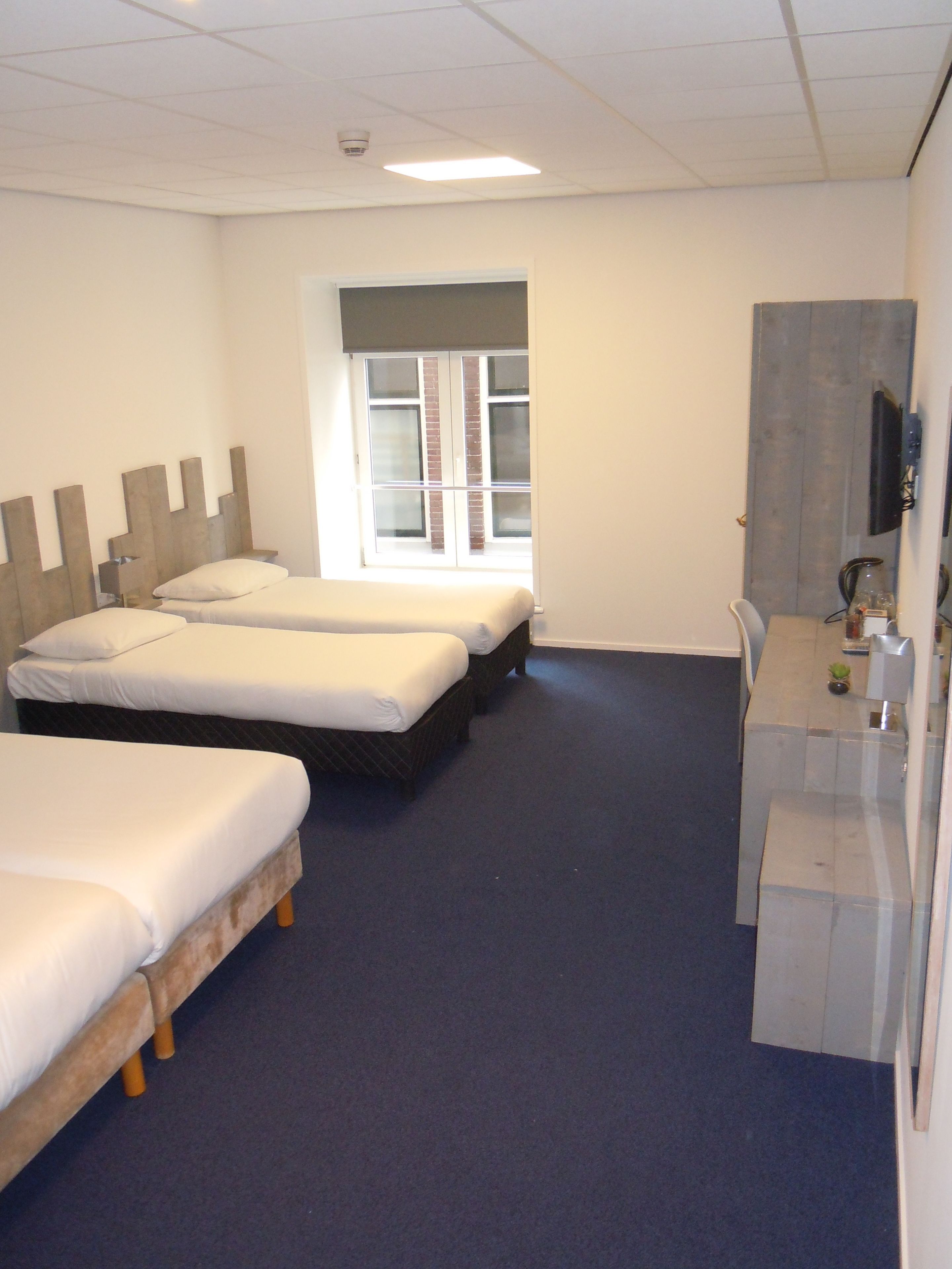 A hotel room with three single beds, a desk with a chair, a television, and a window with blinds.