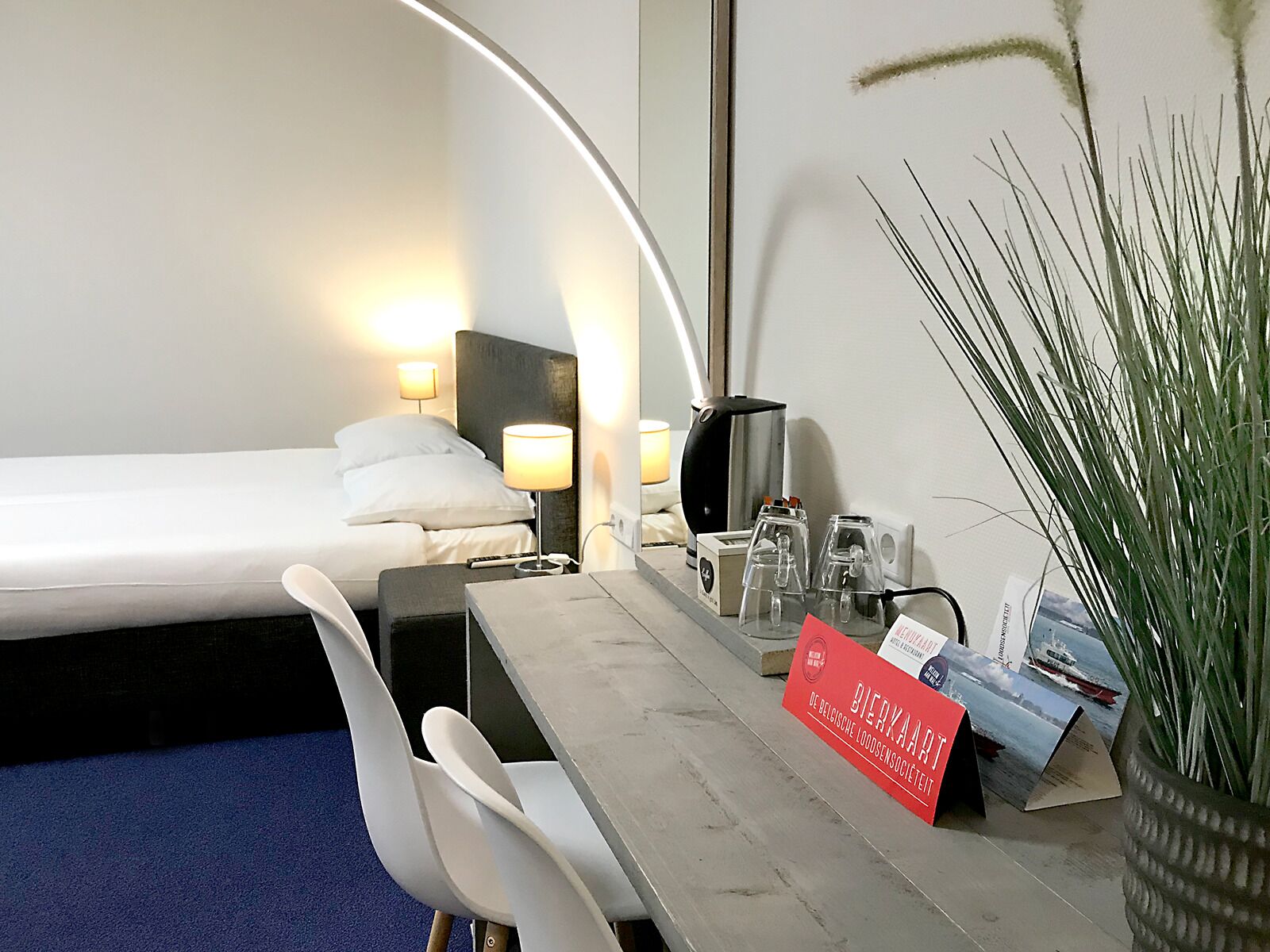 A hotel room with a bed, bedside table, lamp, and a desk with a chair. There is a potted plant and a sign on the desk.