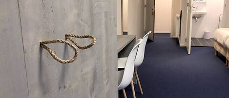Camera d'albergo con porta in legno, una scrivania con due sedie bianche, un letto, un bagno con lavandino e specchio, e un tappeto blu.