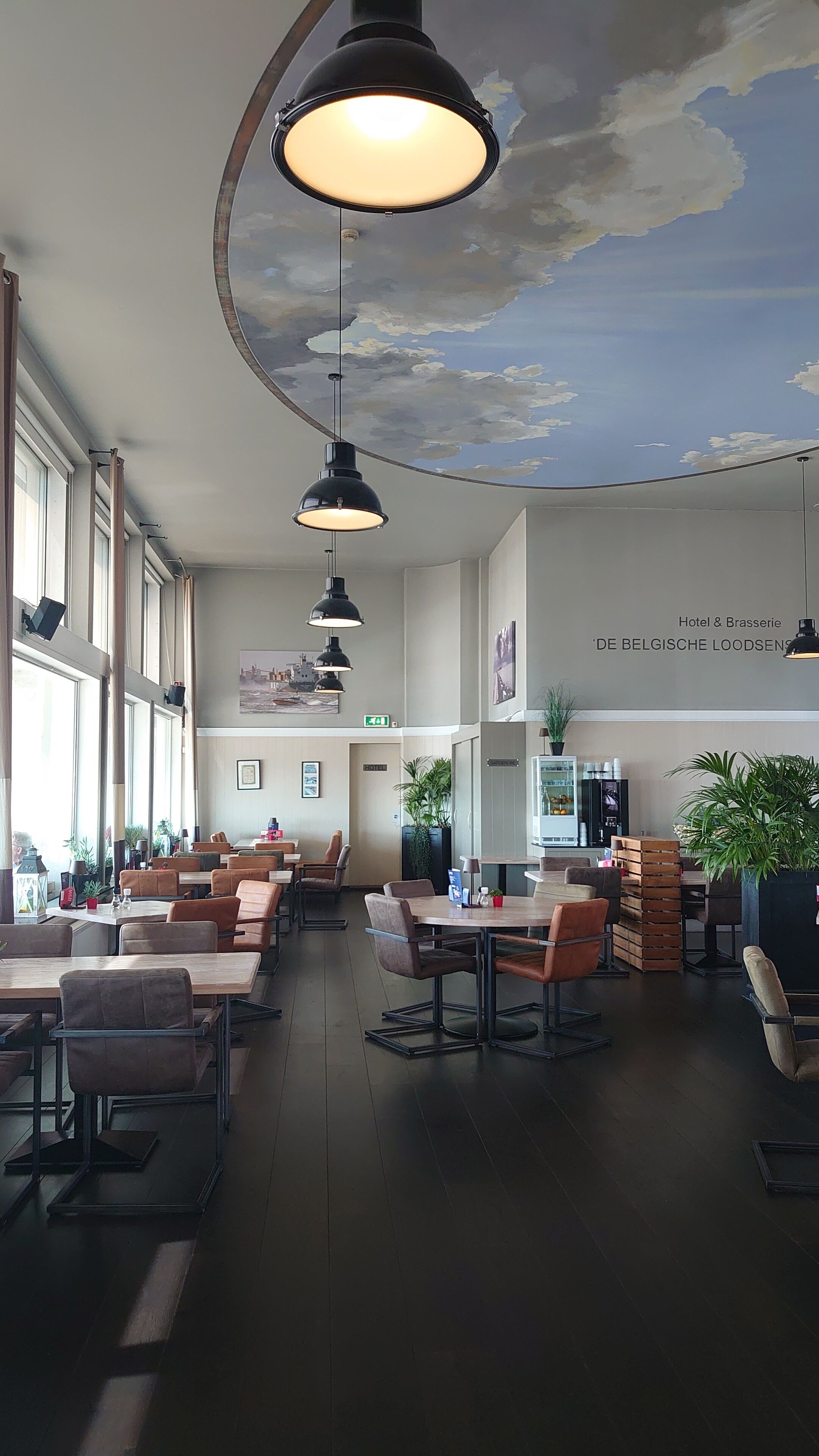 A spacious hotel lobby with high ceilings, large windows, and a mural of clouds. There are multiple seating areas with tables and chairs, and a refrigerator stocked with beverages.