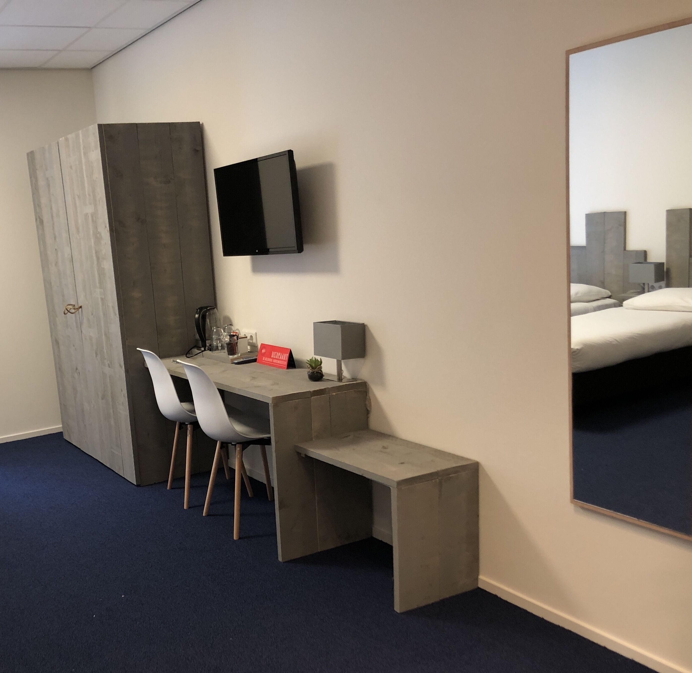 A hotel room with a desk, two chairs, a flat-screen TV, a bed, and a mirror.