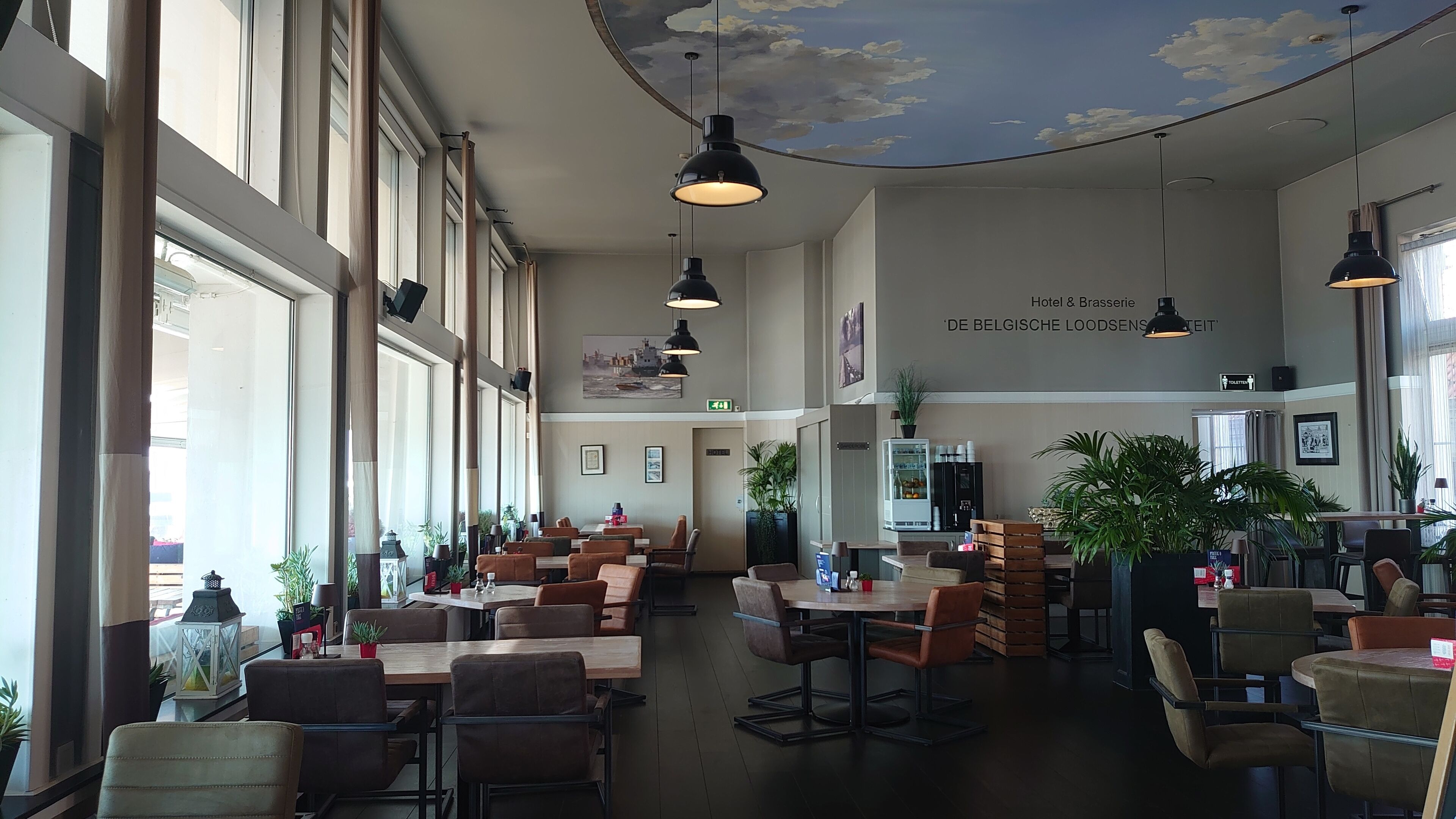 A spacious hotel dining area with high ceilings, large windows, and modern pendant lighting.