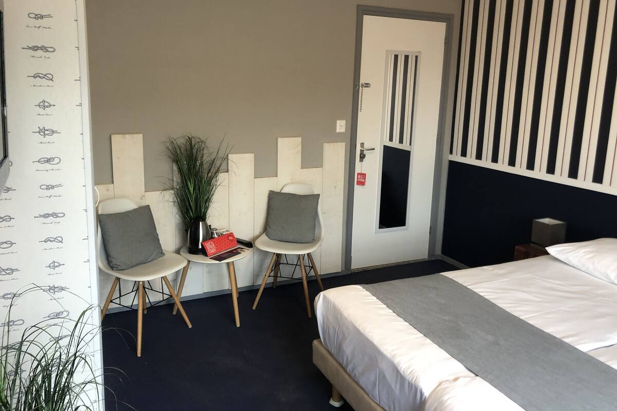 Une chambre d’hôtel avec un lit, deux chaises, une petite table sur laquelle se trouve un objet rouge, une plante en pot et une porte avec une pancarte.
