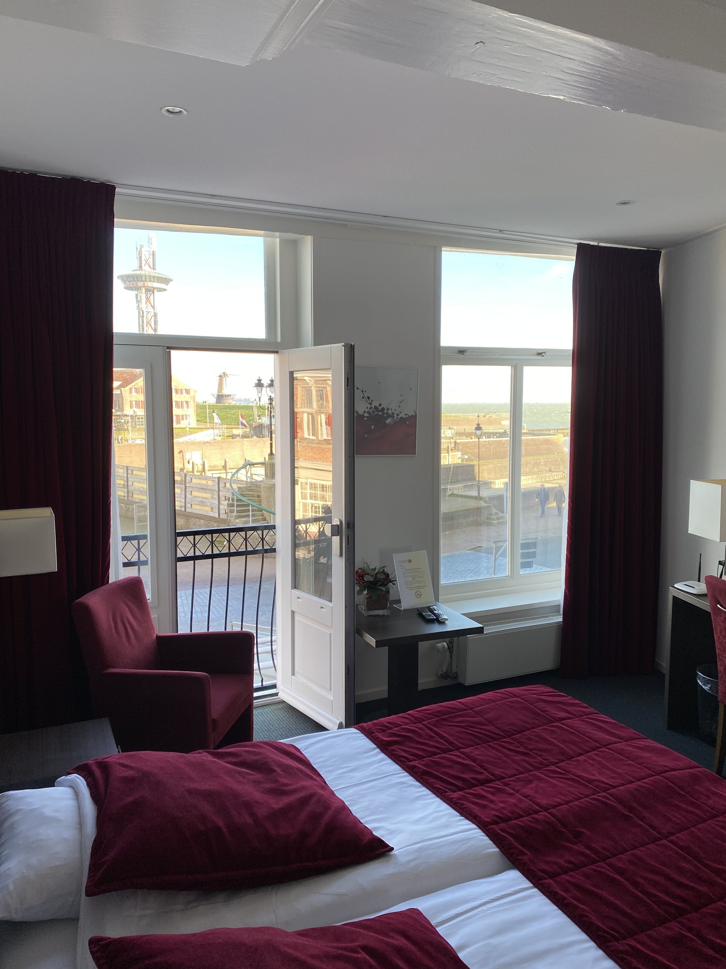 A hotel room with a bed, a desk, a chair, and a view of a cityscape through large windows.