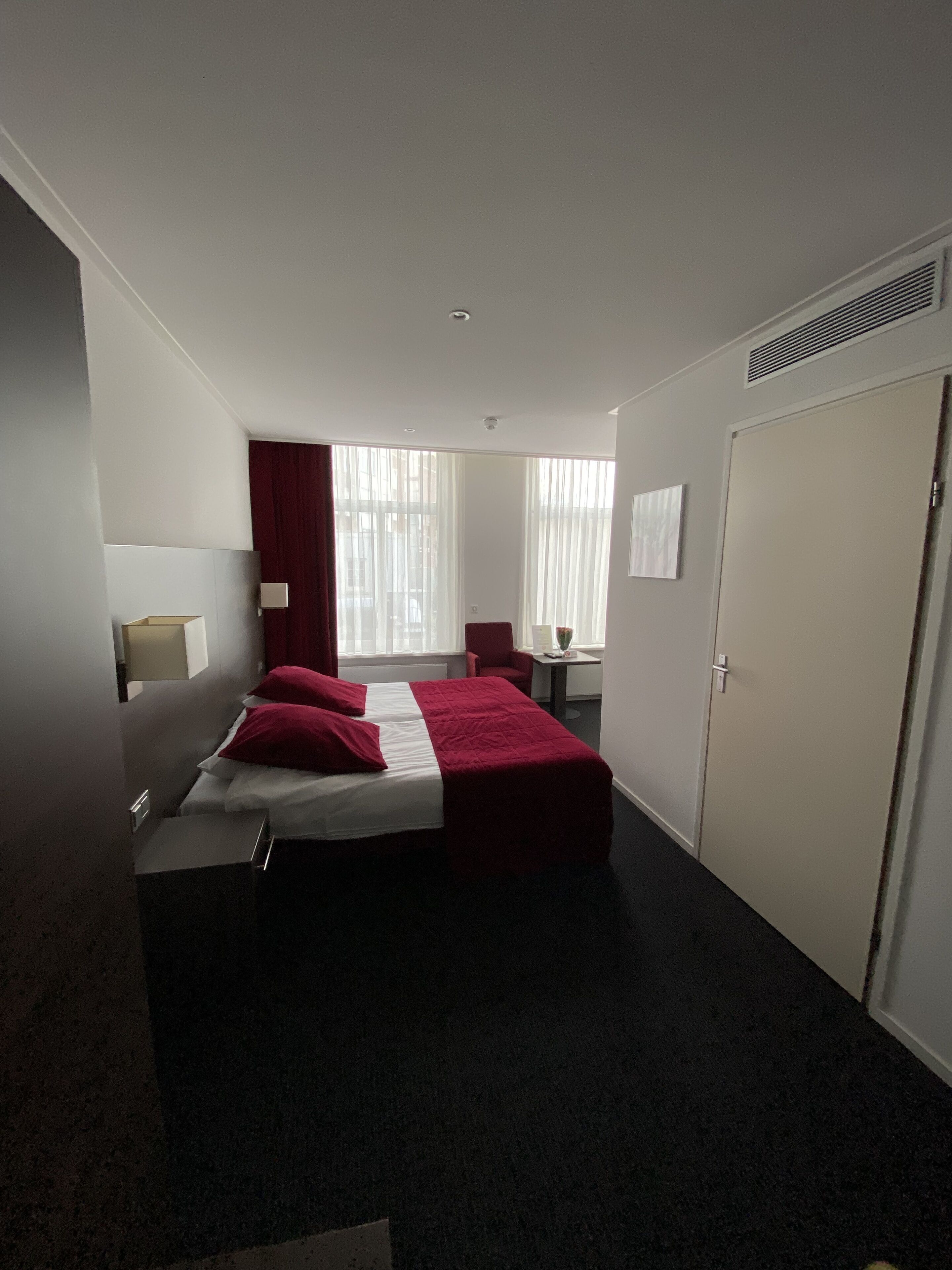 Une chambre d’hôtel comprenant un lit, un bureau, une chaise, une armoire et une fenêtre avec des rideaux.