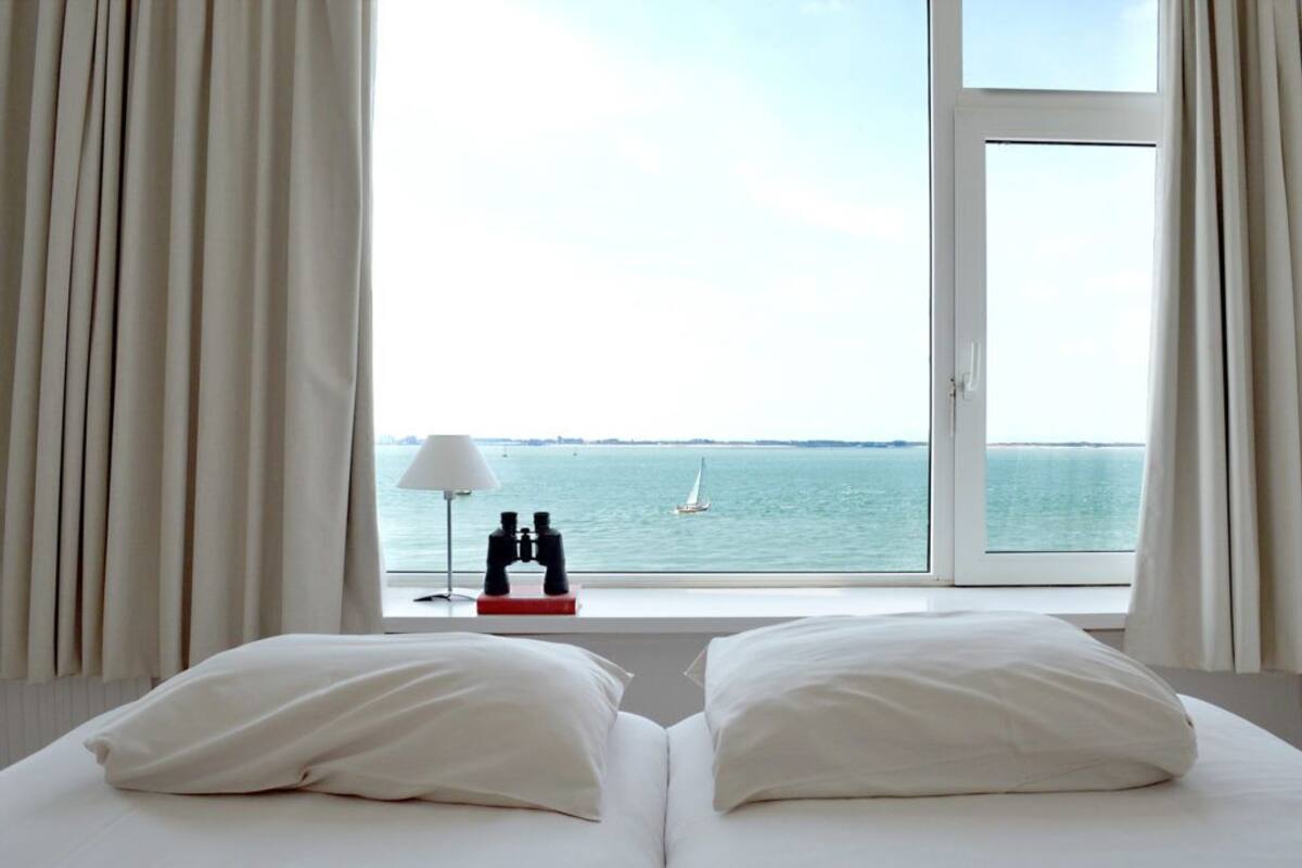 Une chambre d’hôtel avec un lit, une fenêtre avec des rideaux, une lampe et une paire de jumelles posées sur une table.