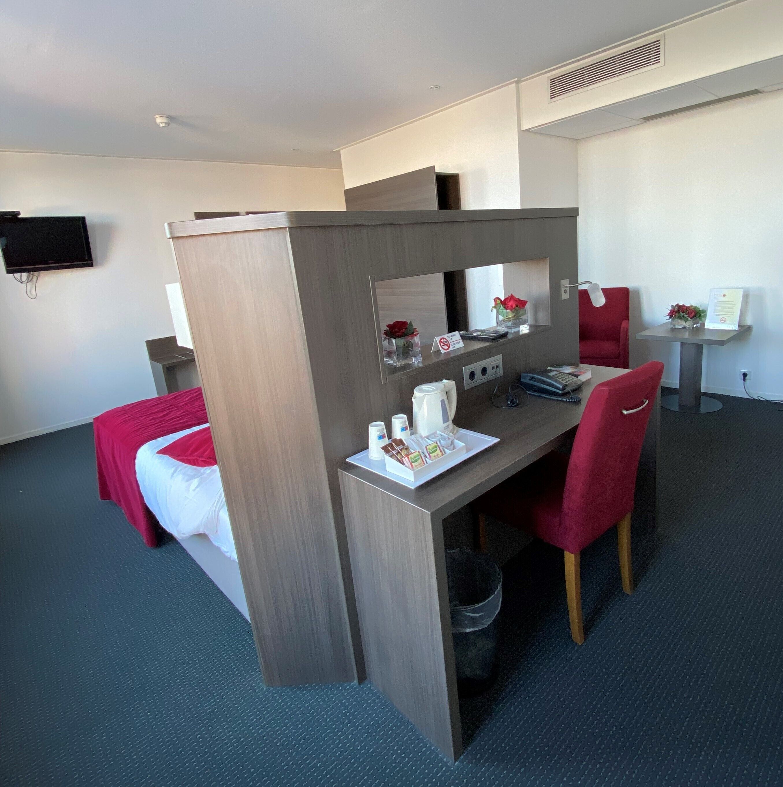 Habitación de hotel con una cama individual, un escritorio con silla, una mesita con un florero y un televisor montado en la pared.