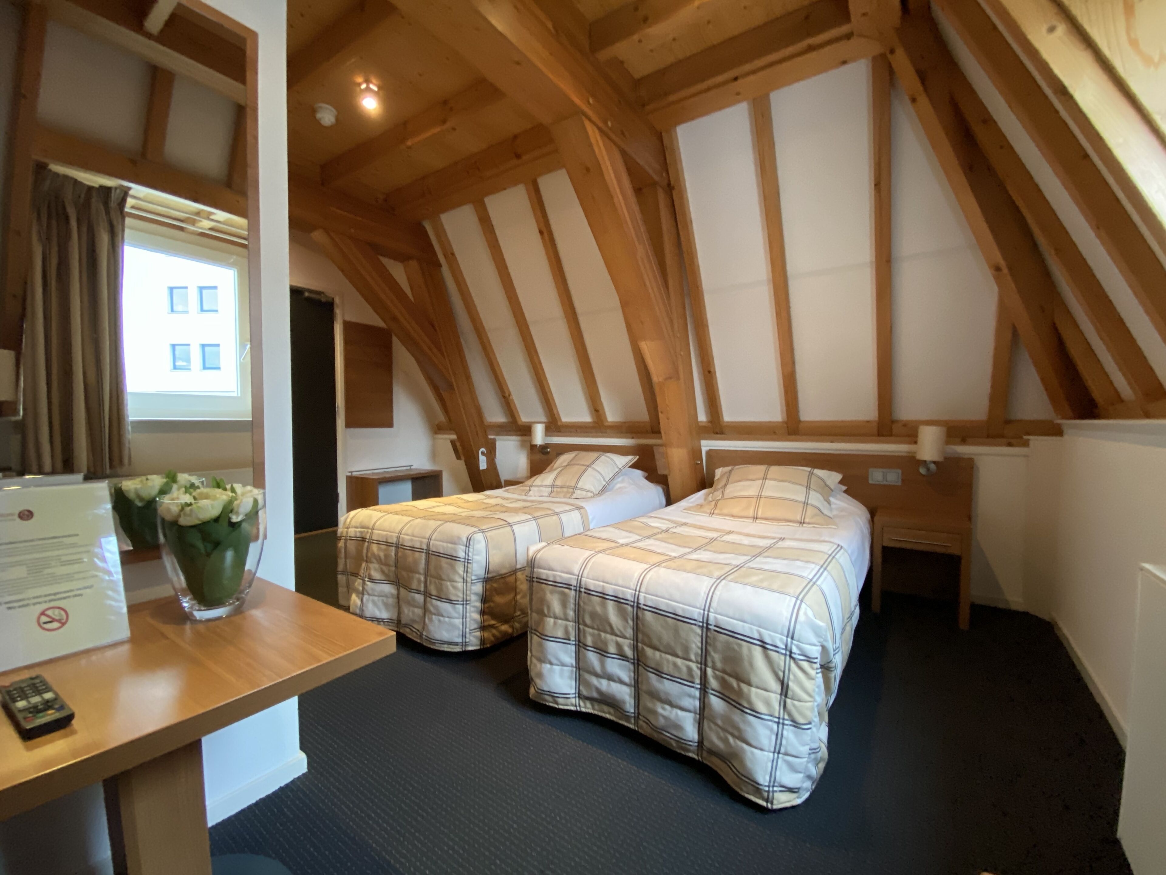 A room with two beds, wooden beams, a desk, a vase with flowers, and a window.