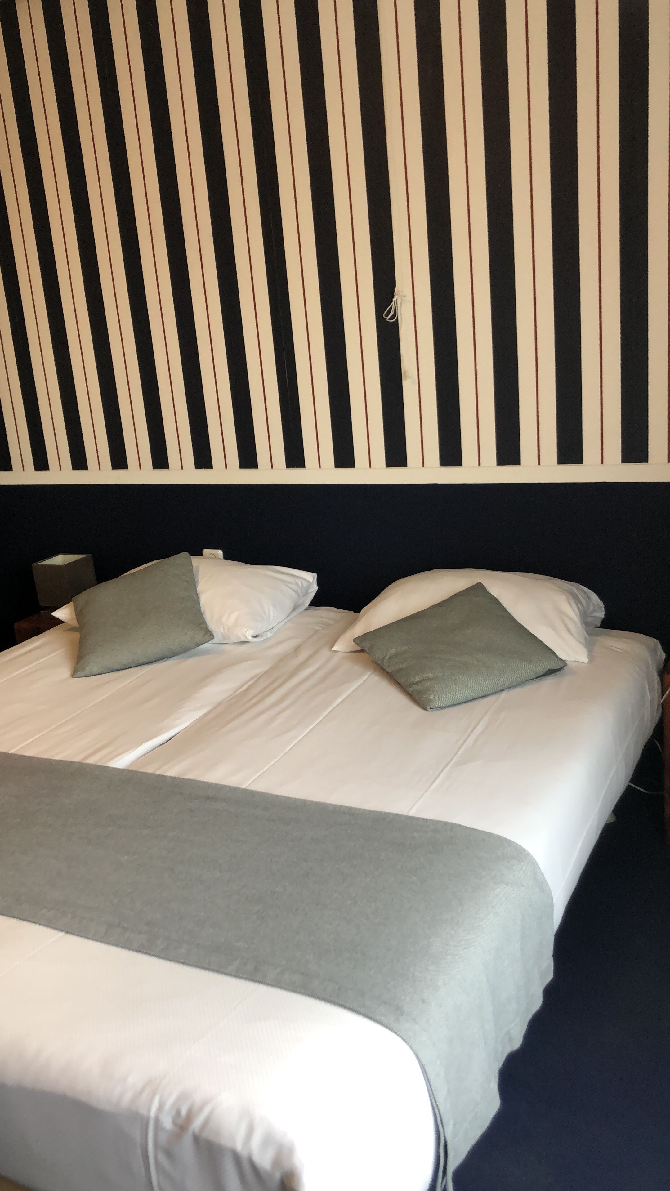 Una habitación de hotel con una cama grande, dos almohadas, una mesita de noche y una pared con rayas verticales.
