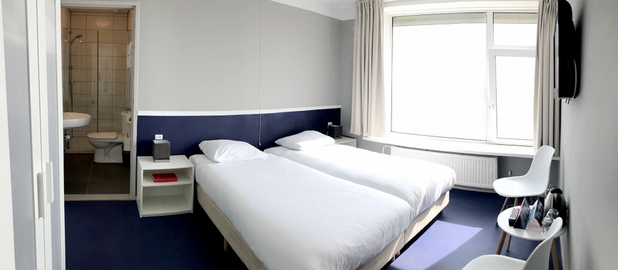 A modern hotel room with two single beds, a bedside table with a lamp, a small nightstand, a window with curtains, and a view of the bathroom.