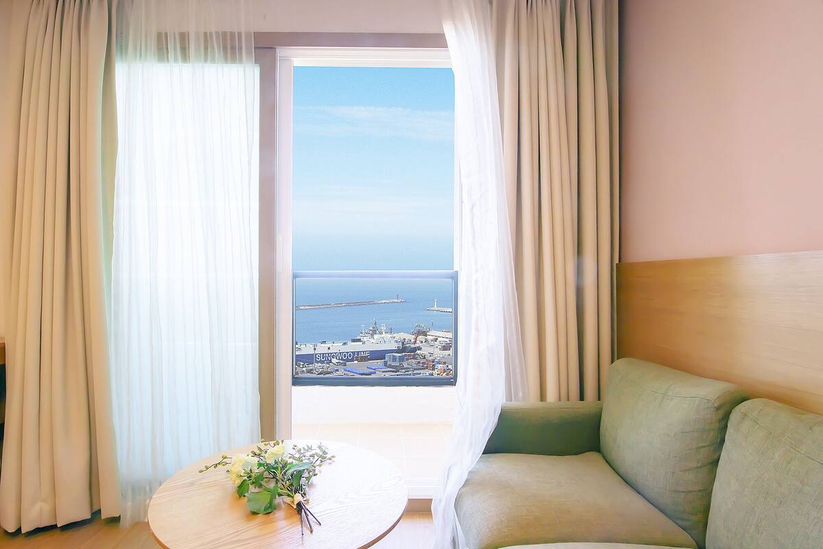 A hotel room with a view of a port and water. There is a green sofa and a small table with a flower arrangement.