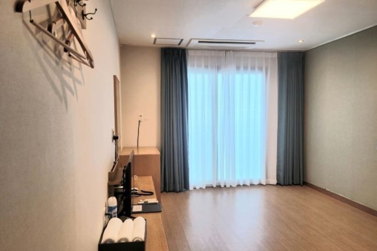 A hotel room with a wooden floor, a desk with a computer, a chair, and a TV. There is a window with curtains and a view of the outside.