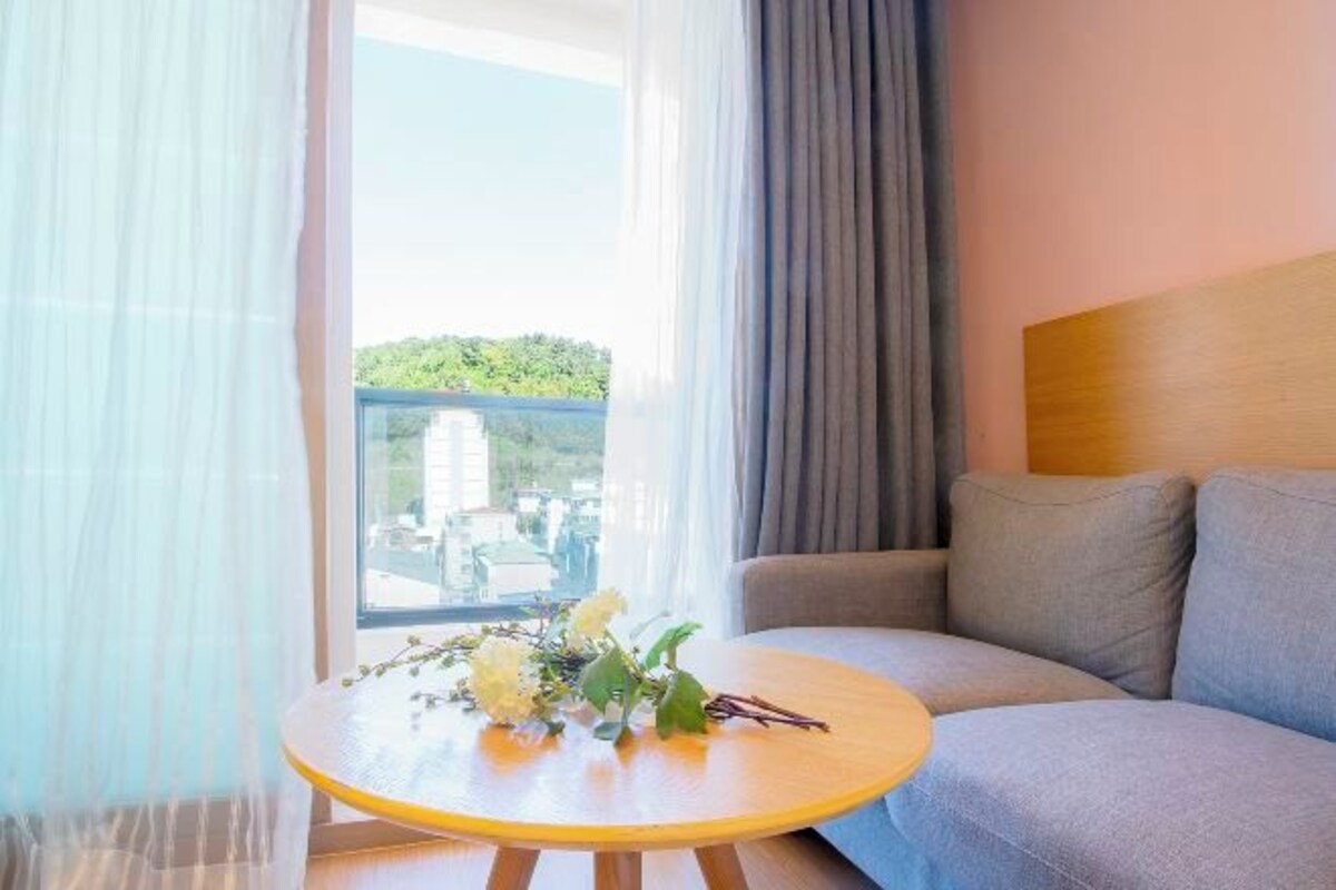 A room with a grey sofa, a small round table with a bouquet of flowers, and a view of greenery and buildings through sheer curtains.