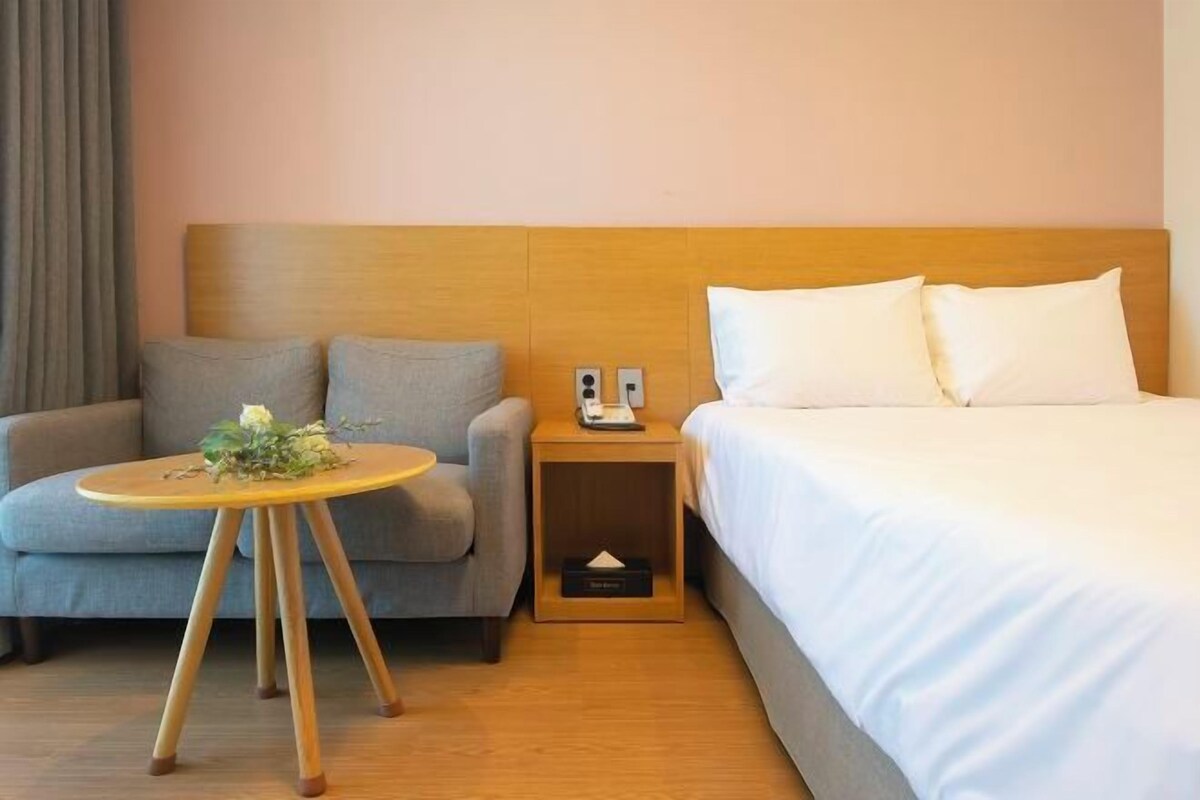 A hotel room with a bed, a sofa, a small wooden nightstand, and a round table with a flower arrangement.