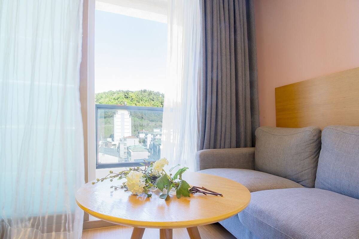 A modern hotel room with a balcony, a round wooden table with a vase of flowers, and a grey sofa.