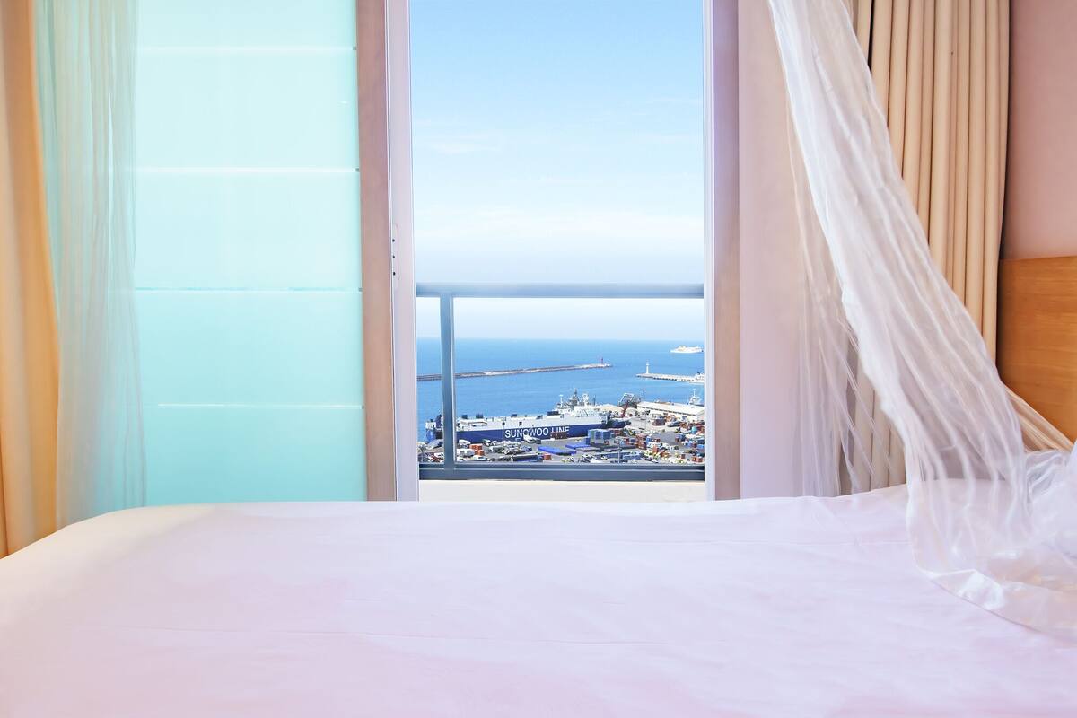 A hotel room with a bed, a view of the sea, and a ship named SUNWOO LINE in the background.