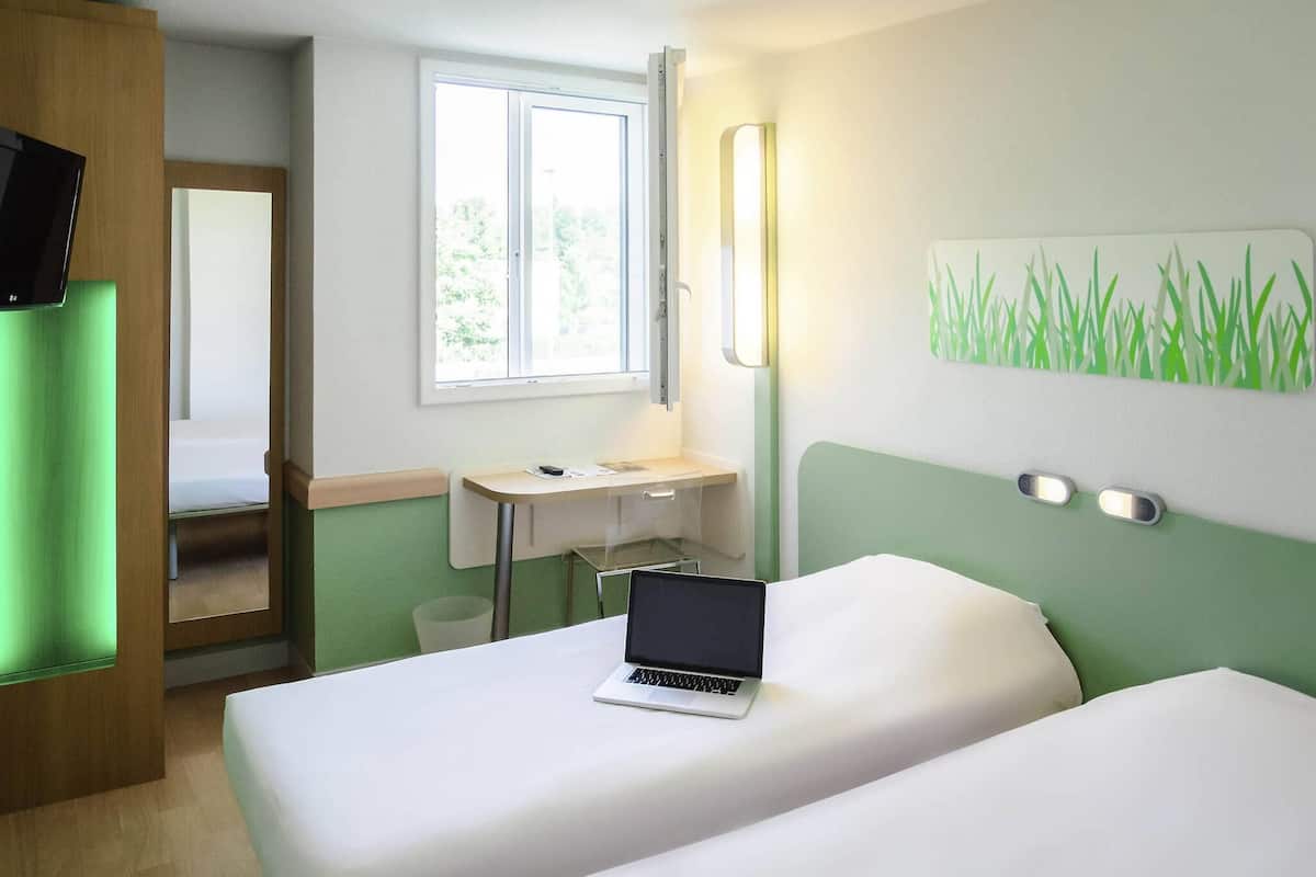 A hotel room with two beds, a desk, a laptop, a window, and a green wall.