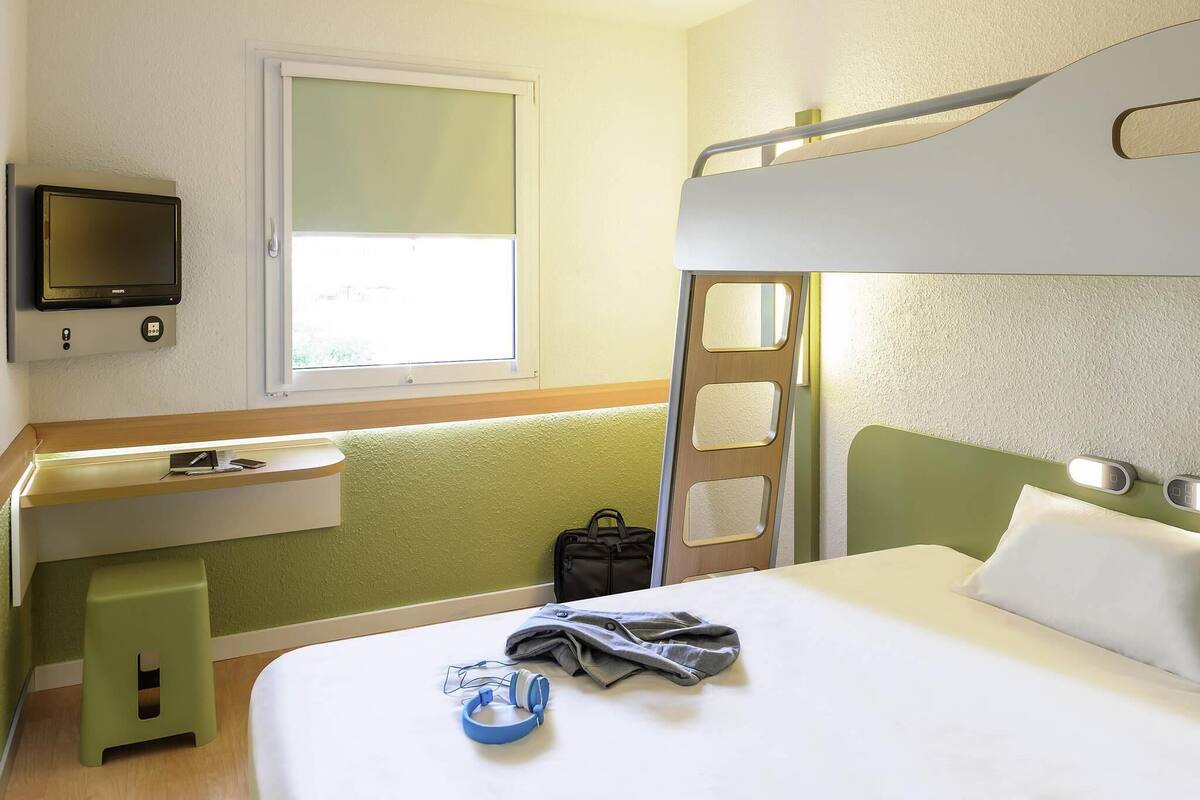 A hotel room with a bunk bed, a desk with a chair, a television mounted on the wall, and a window providing natural light.