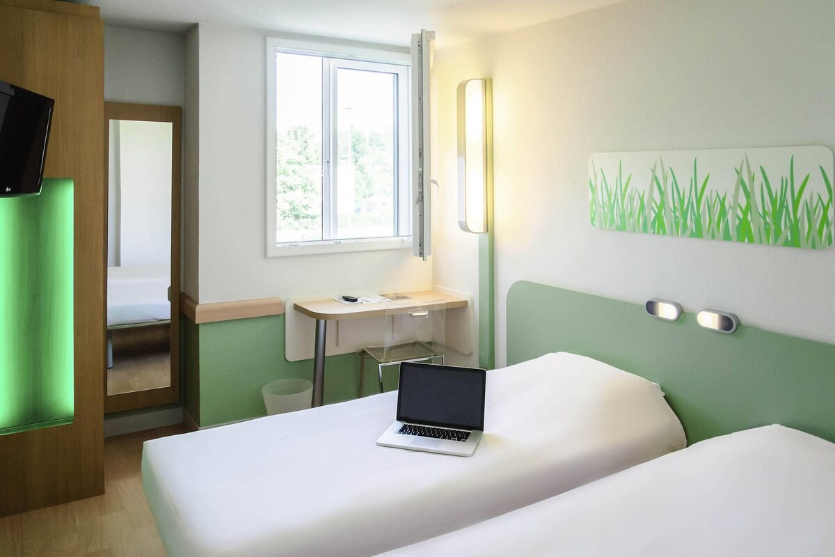 Habitación de hotel con dos camas, un escritorio, una computadora portátil, un ventanal y una pared verde.