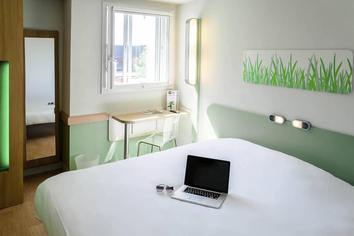 A hotel room with a bed, a desk with a laptop, a window, and a wall-mounted light.