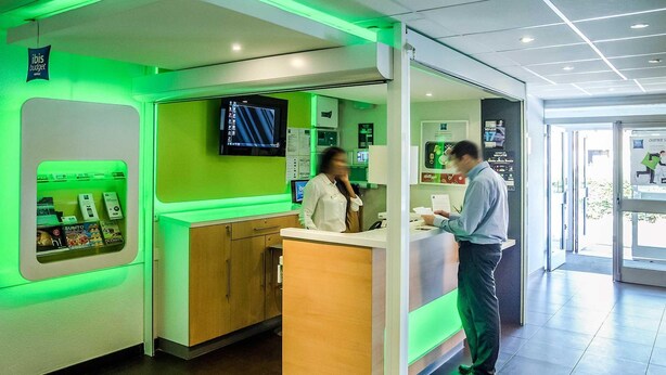 Zona de recepción de hotel con mostrador iluminado en verde, un televisor montado en la pared y un minibar a la vista.
