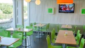 A restaurant interior with green chairs and tables, a large orange overhead light, and a TV on the wall. - ibis budget Aeroport le Bourget Garonor (Le Blanc-Mesnil)