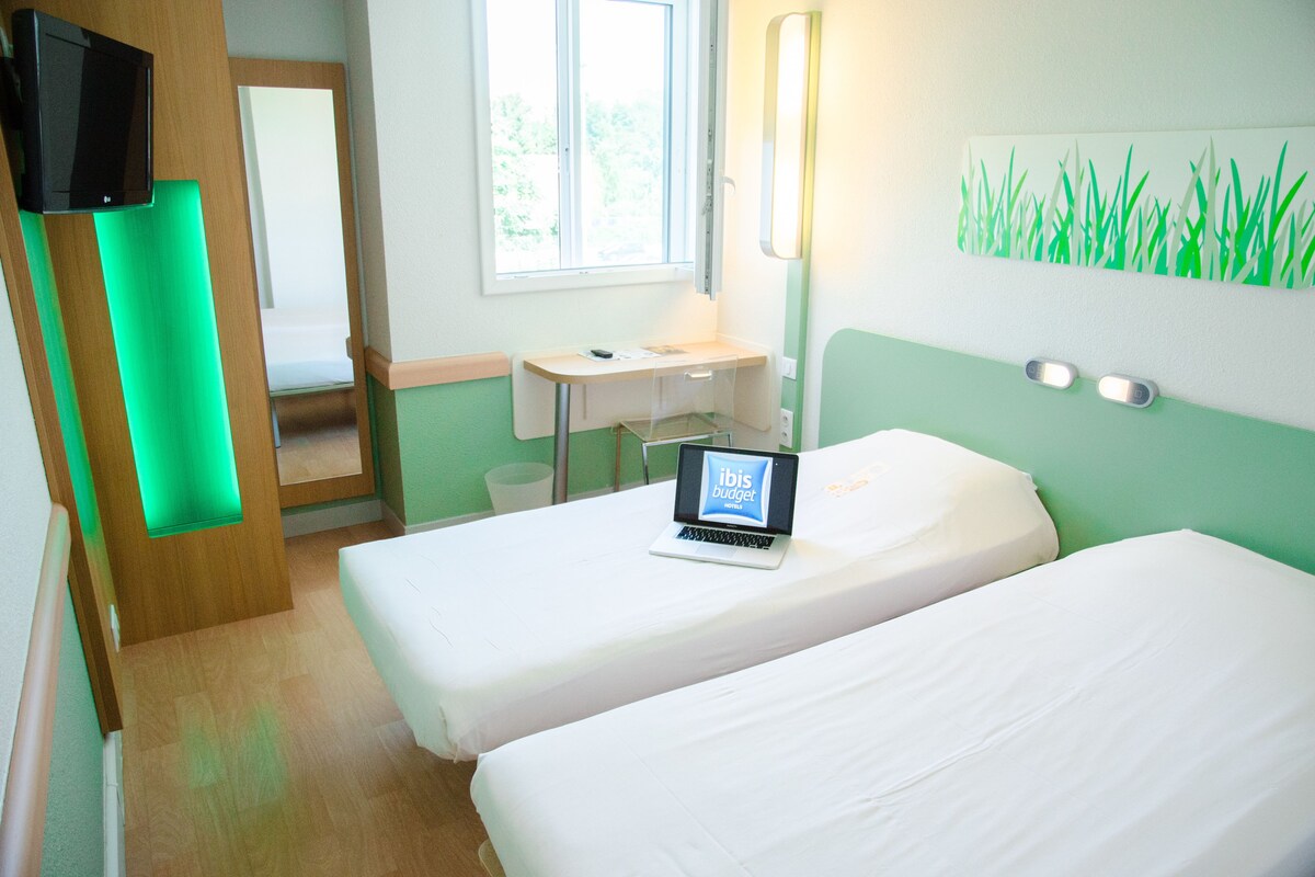 Habitación de hotel con dos camas, un escritorio con una computadora portátil, un televisor y una ventana con vista a follaje.
