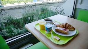 Bandeja de desayuno con croissant, jugo, café y yogur sobre una mesa junto a una ventana con vistas a follaje y una cerca.