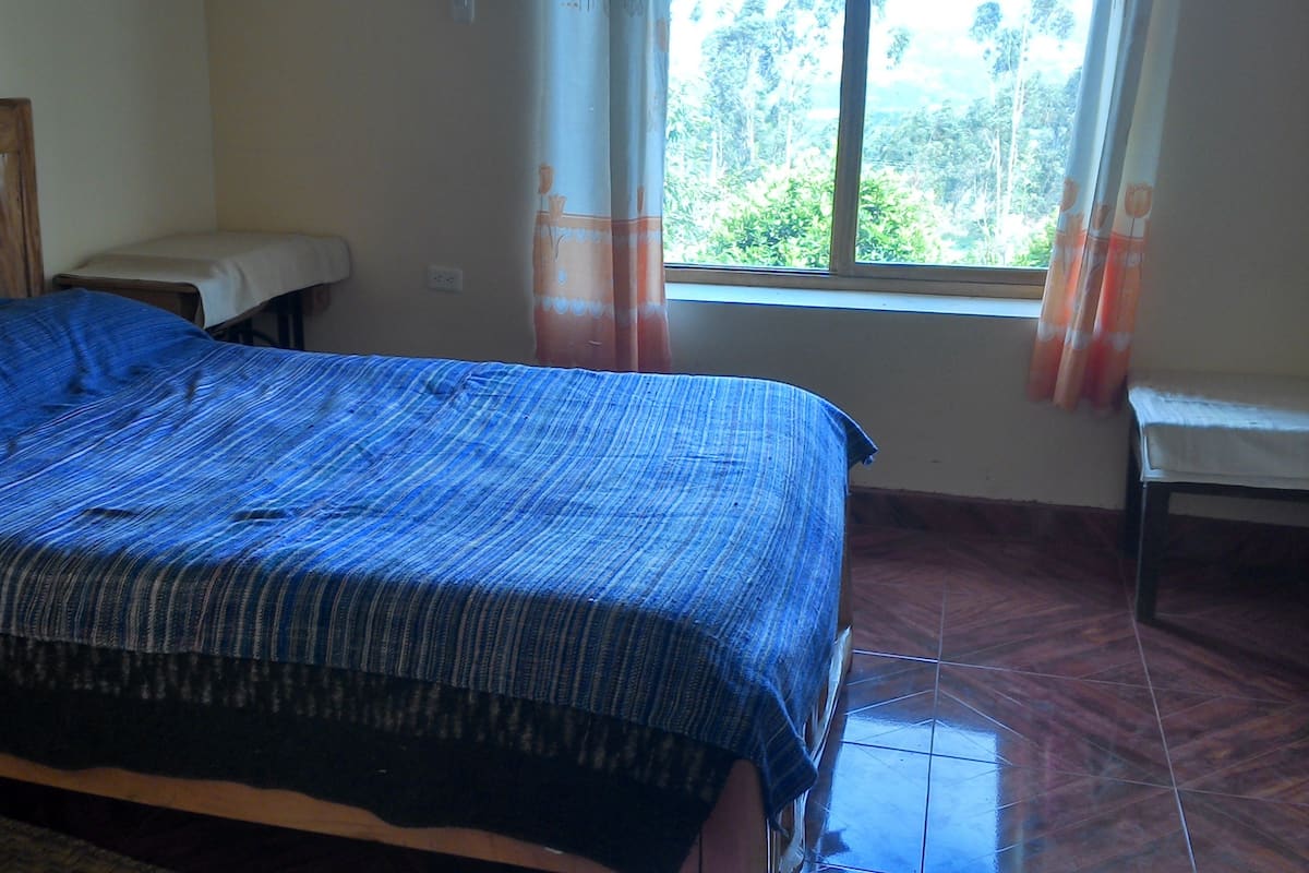 A bedroom with a bed, a wooden headboard, a bench, a window with curtains, and a tiled floor.