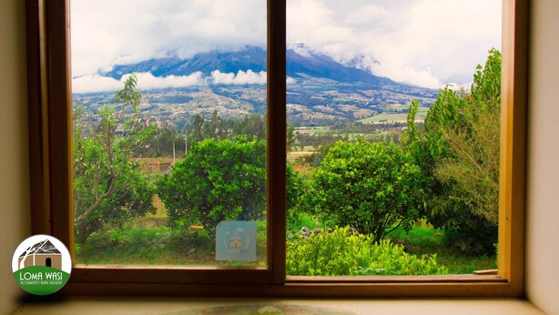 A window view of a mountainous landscape with a logo of Loma Wasi in the bottom left corner. - Loma Wasi Village (Cotacachi)