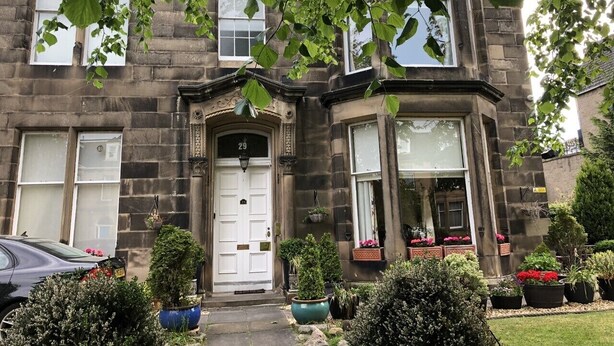 A two-story stone building with a white front door, multiple windows, and a small garden with potted plants and flowers. - The Alba House (Edinburgh)