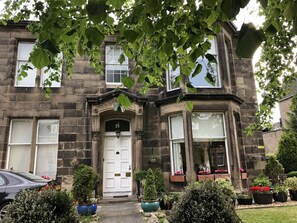 A two-story stone building with a white front door, multiple windows, and a small garden with potted plants and flowers. - The Alba House (Edinburgh)