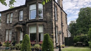 A stone building with a large bay window, a front door, and a small garden with potted plants and a lawn. - The Alba House (Edinburgh)