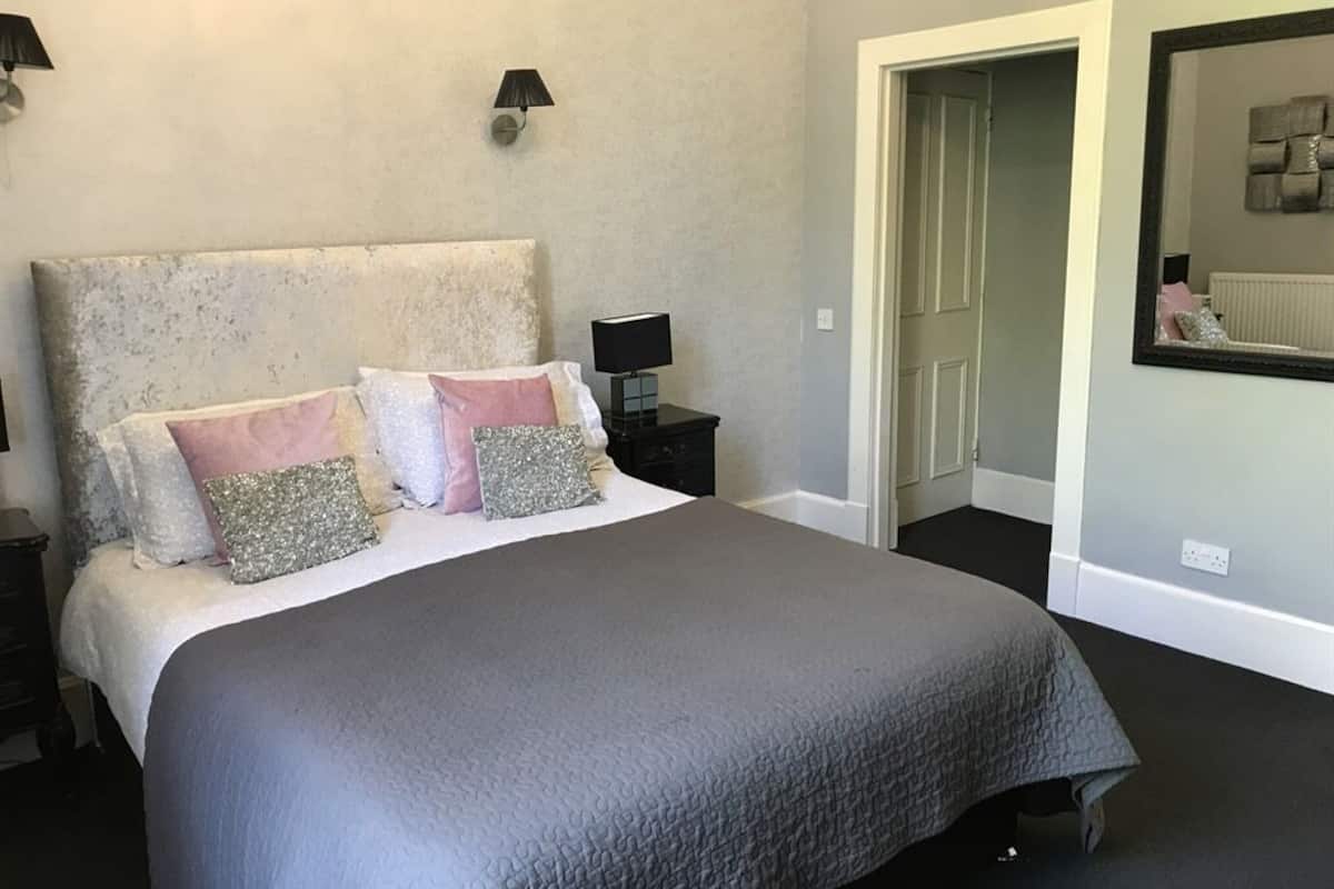 A bedroom with a large bed, a nightstand with a lamp, a door, and a mirror on the wall.